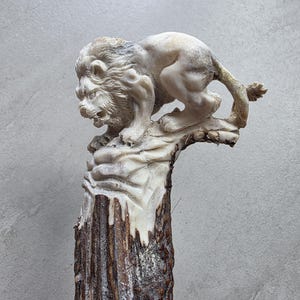 Peut inclure: Une sculpture détaillée d'un lion en position accroupie, fabriquée à partir d'un matériau de couleur claire. La crinière et les traits du visage du lion sont finement détaillés. La sculpture est montée sur une base texturée brun foncé, peut-être un morceau de bois ou de bois de cerf.