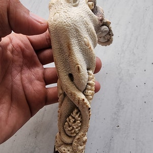 May include: A carved octopus sculpture in shades of cream and brown. The octopus is intricately detailed with textured skin and curled tentacles. The sculpture is held in a hand, showcasing the detailed craftsmanship of the marine-themed art piece.