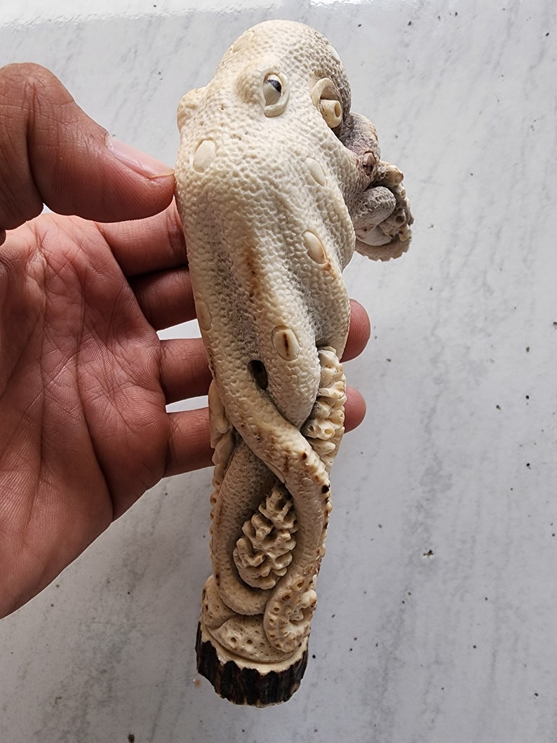 May include: A carved octopus sculpture in shades of cream and brown. The octopus is intricately detailed with textured skin and curled tentacles. The sculpture is held in a hand, showcasing the detailed craftsmanship of the marine-themed art piece.