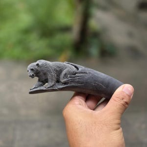 May include: A dark grey, carved wooden sculpture of a bear lying on a log. The bear is detailed with fur and facial features. The sculpture is held in a hand against a blurred green background.