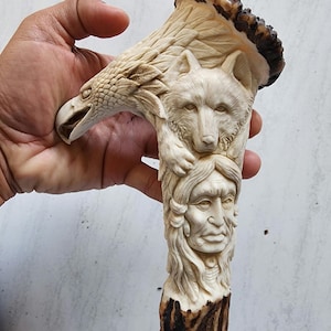 Peut inclure: Une sculpture en corne sculptée représentant un aigle, un loup et un visage amérindien. La sculpture est de couleur crème avec des accents bruns et des détails complexes. L'aigle est en haut, le loup au milieu et le visage en bas.