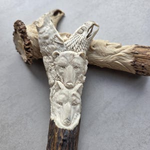 May include: A carved antler sculpture featuring a wolf, bear, and eagle. The sculpture is cream-colored with detailed carvings of the animals' faces. The base of the antler is a dark brown color. The sculpture is a unique piece of art.