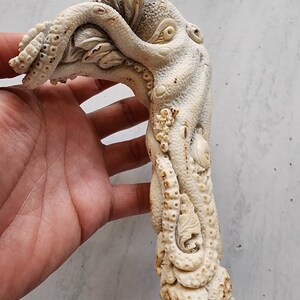 May include: A detailed, ivory-colored sculpture of an octopus, carved with intricate details. The octopus's tentacles wrap around a central form, with a textured surface and visible suckers. The base is a dark brown, possibly wood or antler.