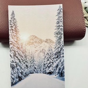 May include: A rectangular card featuring a winter landscape. The image shows a snow-covered road winding through a forest of snow-laden evergreen trees, leading towards snow-capped mountains under a bright sky.