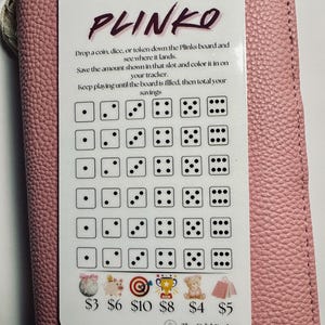 May include: A white Plinko savings tracker card with black dice patterns and the word "PLINKO" in purple. The card includes instructions and savings amounts from $3 to $10. The card is on a pink textured surface.