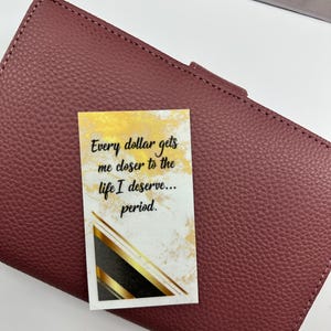 May include: A burgundy leather wallet with a textured surface. A small card with gold accents and the text "Every dollar gets me closer to the life I deserve... period." is on top of the wallet.