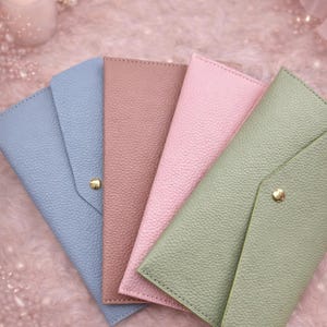 May include: Four pastel-coloured wallets arranged on a pink, textured surface. The wallets are in shades of blue, mauve, pink, and green. Each wallet has a textured finish and a gold-coloured button closure.