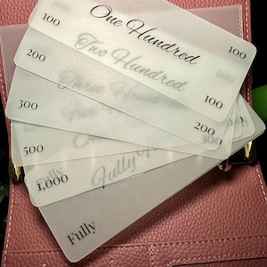 Puede incluir: Un conjunto de tarjetas translúcidas con valores de 100, 200, 300, 500 y 1.000, y las palabras "One Hundred" y "Two Hundred" en escritura cursiva. Las tarjetas están dispuestas sobre un planificador de cuero rosa.