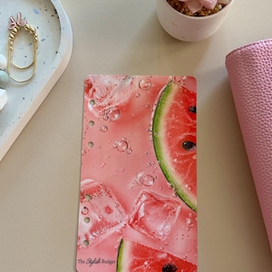 Op de afbeelding: Een rechthoekig planner-dashboard met een watermeloen- en ijsblokjesafbeelding op een roze achtergrond met bubbels. De linkerkant heeft zes ponsgaten voor in een map, en de tekst The Stylish Budget staat linksonder afgedrukt.