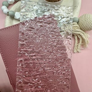 May include: A clear, textured glass rectangle with a bubbly pattern. The glass is displayed over a pink leather surface. A small succulent in a white pot and decorative beads are in the background.