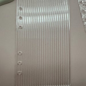 May include: A clear, rectangular sheet of ridged plastic with four circular holes along the left edge. The sheet has vertical lines running from top to bottom. The object is transparent and appears to be a planner insert.