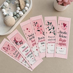 May include: A set of five rectangular pink bookmarks with vertical stripes, floral and celestial illustrations, and motivational text including "Breathe. Release. Begin again." and "Rest is Productivity too." Each bookmark features punched holes.