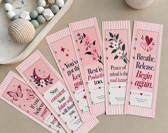 May include: A set of five rectangular pink bookmarks with vertical stripes, floral and celestial illustrations, and motivational text including "Breathe. Release. Begin again." and "Rest is Productivity too." Each bookmark features punched holes.