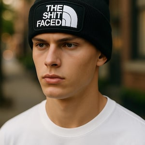 May include: Black beanie hat with a white rectangular patch displaying the text "THE SHIT FACED". The hat is worn by a person wearing a white t-shirt. The beanie is a casual accessory.