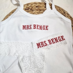 May include: White tank top and matching underwear set with the embroidered text "MRS. BENGE" in red. The underwear features a white lace trim. The tank top has thin straps.