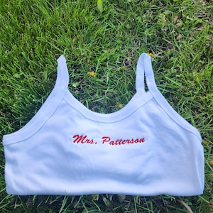 May include: A white tank top with thin straps, featuring the embroidered text "Mrs. Patterson" in red. The top is made of a soft material and is designed for comfort. The garment is displayed on a bed of green grass.