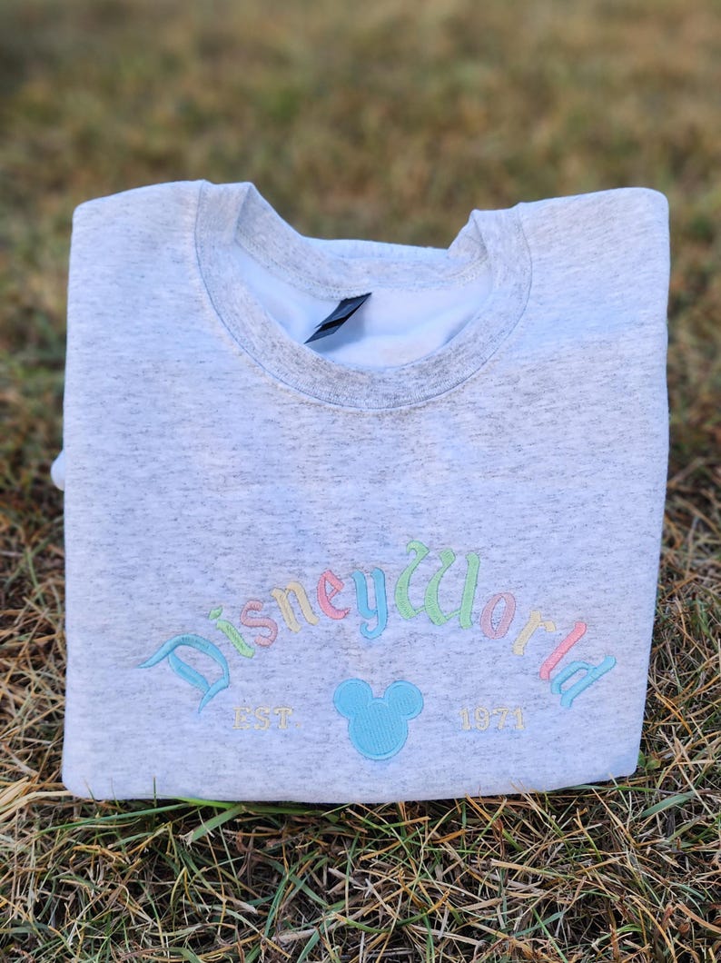 May include: A gray sweatshirt with a rainbow-colored embroidered "Disney World" logo, a blue Mickey Mouse silhouette, and the text "EST 1971".