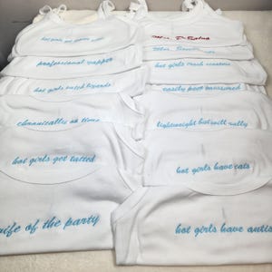 May include: A collection of white tank tops featuring various embroidered phrases in light blue thread, including "professional yapper," "hot girls catch lizards," "hot girls get tatted," "life of the party," "hot girls have cats," and "hot girls have autism."