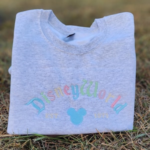 May include: Gray sweatshirt with a rainbow colored "Disney World" logo and a blue Mickey Mouse head. The text "EST 1971" is below the logo.
