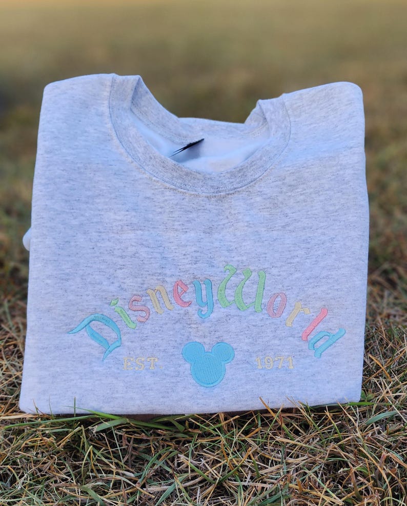 May include: Gray sweatshirt with a rainbow colored "Disney World" logo and a blue Mickey Mouse head. The text "EST 1971" is below the logo.