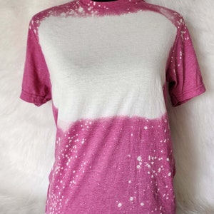 Puede incluir: Una camiseta rosa y blanca con un dise&ntilde;o salpicado de lej&iacute;a. La camiseta tiene cuello redondo y mangas cortas.