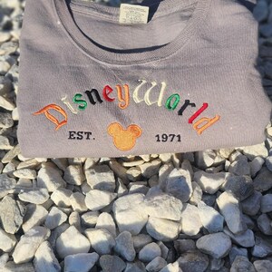 May include: A gray t-shirt with the text "Disneyworld Est. 1971" embroidered in colorful letters. A cartoon Mickey Mouse head is embroidered in orange and yellow next to the text.