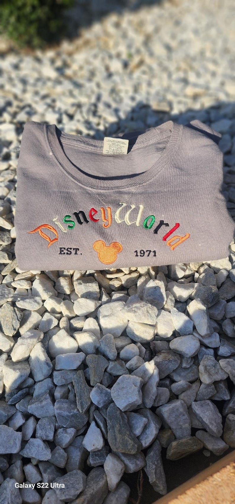 May include: A gray t-shirt with the text "Disneyworld Est. 1971" embroidered in colorful letters. A cartoon Mickey Mouse head is embroidered in orange and yellow next to the text.