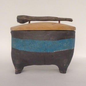 Peut inclure: Une petite boîte en céramique gris foncé avec un couvercle en bois et un morceau de bois flotté pour poignée. La boîte a une bande bleue autour du milieu.