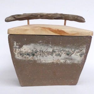 Könnte beinhalten: Eine quadratische Keramikbox mit einem Deckel aus hellem Holz. Der Deckel hat einen Griff aus einem natürlichen Ast. Die Box ist mit einer braunen Glasur mit einem weißen und grauen Design versehen.