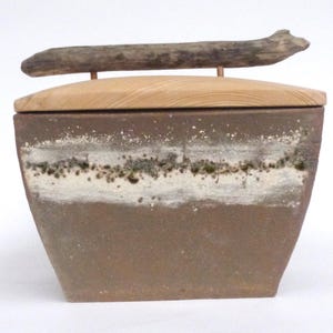Könnte beinhalten: Eine rechteckige Keramikbox mit einem Holzdeckel und einem Treibholzgriff. Die Box hat eine braune und weiße Glasur mit einer strukturierten Oberfläche.