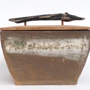 Puede incluir: Una caja de cerámica con tapa y asa de madera. La caja está decorada con un esmalte blanco y marrón y tiene un patrón moteado. El asa está hecha de una pieza natural de madera flotante.