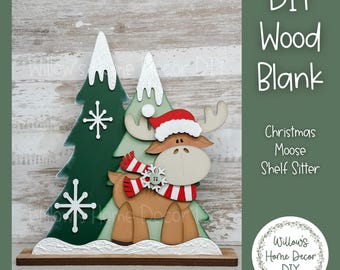 Puede incluir: Adorno navideño de madera con un alce que lleva un gorro de Papá Noel y una bufanda a rayas rojas y blancas. El alce está delante de dos árboles de Navidad verdes con detalles de nieve blanca. El texto "DIY Wood Blank" es visible.