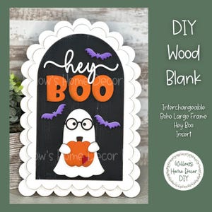 May include: A white scalloped wooden frame with a black background and a ghost holding a pumpkin. The text "hey BOO" is written in orange and black letters on the frame.