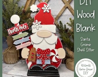 Puede incluir: Un gnomo de Santa de madera DIY. El gnomo tiene un gorro rojo con copos de nieve blancos, barba blanca y nariz beige. Sostiene un regalo, un hombre de jengibre y un bastón de caramelo. Un letrero apunta al Polo Norte, el taller de Santa Claus, y la aldea de los elfos.