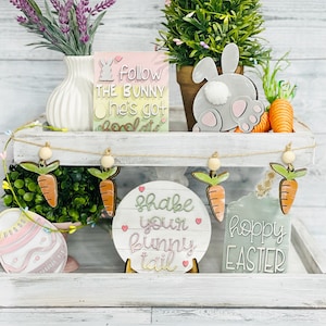 May include: A tiered white shelf with a wooden top and bottom. The top shelf has a small green plant, a gray bunny cutout, and a sign that says "Follow the bunny he's got chocolate". The bottom shelf has a green plant, a sign that says "Shake your bunny tail", a sign that says "Hoppy Easter", and a pink and white Easter egg cutout.