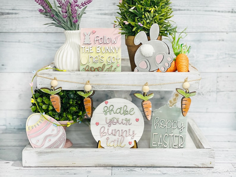 May include: A tiered white shelf with a wooden top and bottom. The top shelf has a small green plant, a gray bunny cutout, and a sign that says "Follow the bunny he's got chocolate". The bottom shelf has a green plant, a sign that says "Shake your bunny tail", a sign that says "Hoppy Easter", and a pink and white Easter egg cutout.