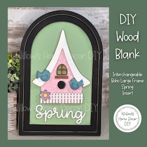May include: A wooden blank sign with a black frame and a green background. The sign features a pink birdhouse with a white roof, two blue birds, and a white picket fence. The word "Spring" is written in black letters on the sign. The sign is labeled "Interchangeable Boho Large Frame Spring Insert".