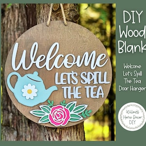 May include: A round wooden door hanger with the words "Welcome, Let's Spill The Tea" in white script. It features a light blue teapot, a pink rose, and green leaves. The sign is hanging from a tree.