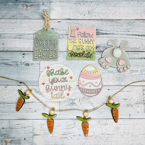 May include: A set of Easter-themed wooden decorations, including a bunny butt, an egg, a tag that says "Hoppy Easter", a round sign that says "Shake your bunny tail", and a tag that says "Follow the bunny he's got chocolate". There is also a garland of carrots with wooden beads.