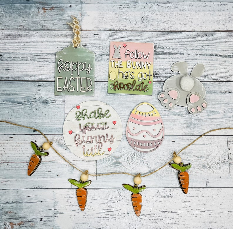 May include: A set of Easter-themed wooden decorations, including a bunny butt, an egg, a tag that says "Hoppy Easter", a round sign that says "Shake your bunny tail", and a tag that says "Follow the bunny he's got chocolate". There is also a garland of carrots with wooden beads.