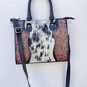 May include: A black and white cowhide tote bag with a black leather handle and shoulder strap. The bag features brown floral tooled leather accents with turquoise and rhinestone embellishments.