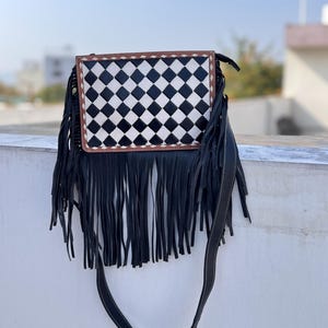 May include: A black and white checkered crossbody bag with long black fringe. The bag has a brown border and a black strap. The bag is made of leather and has a unique design.