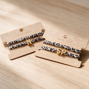 Könnte beinhalten: Zwei Sets von Haargummis mit Leopardenmuster auf beigefarbenen Pappkarten. Jedes Set enthält einen goldfarbenen Metallbuchstaben-Anhänger, wobei ein Set den Buchstaben N und das andere die Buchstaben SP zeigt. Die Karten sind mit dem Text WITH LOVE bedruckt.