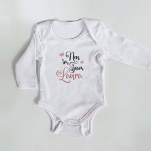 Könnte beinhalten: Weißer Langarm-Babybody mit dem Text "New in Team Laura" in Schwarz und rosa Herzen.
