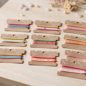 Könnte beinhalten: Eine Sammlung von elastischen Haarbändern oder Armbändern auf braunen Pappkarten. Jede Karte zeigt farbenfrohe Stoffbänder mit aufgedruckten Wörtern wie LOVE, GOOD und BEST FRIENDS sowie Herz- und Smiley-Symbolen.