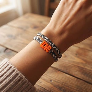 Könnte beinhalten: Doppelreihiges Armband mit einem schwarz-weißen Tiermuster. Das orangefarbene Wort "love" ist auf dem Armband zentriert. Das Armband wird am Handgelenk einer Person getragen.