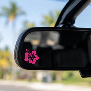 May include: A bright pink hibiscus flower decal applied to the lower left corner of a rectangular vehicle rearview mirror. The floral graphic features distinct petals and a central stamen, contrasting against the reflective glass surface.