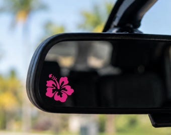 May include: A bright pink hibiscus flower decal applied to the lower left corner of a rectangular vehicle rearview mirror. The floral graphic features distinct petals and a central stamen, contrasting against the reflective glass surface.