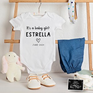 May include: White onesie with the text "It's a baby girl! ESTRELLA" and a heart symbol. The text "JUNE 2024" is below the name. The onesie is on a wooden ladder with a pair of white shoes, a stuffed bunny, a pair of blue denim pants, and a baby ultrasound image.