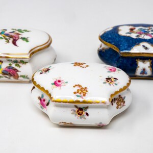 Puede incluir: Tres cajas de porcelana decorativas para alhajas con intrincados diseños florales y de pájaros. Las cajas son en tonos de blanco, azul y dorado.